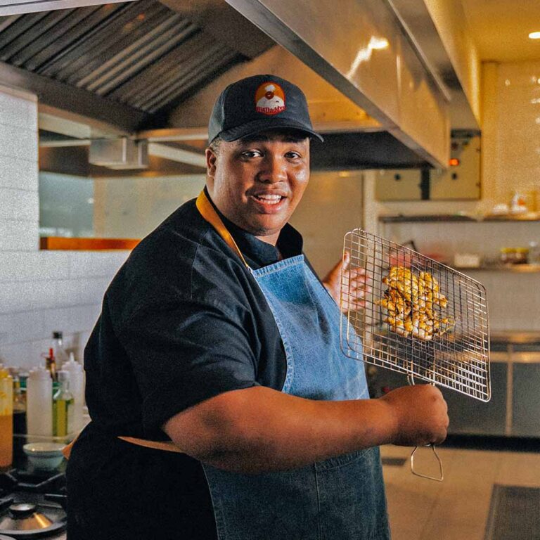 Large black man wearing a denim apron and holding a portable grilling basket near a stove in a commercial kitchen.