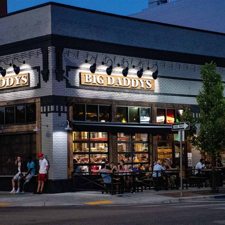 Exterior of busy restaurant on city street corner at night with signs under spotlights that read “Big Daddys”. Customers are eating and loitering outside.