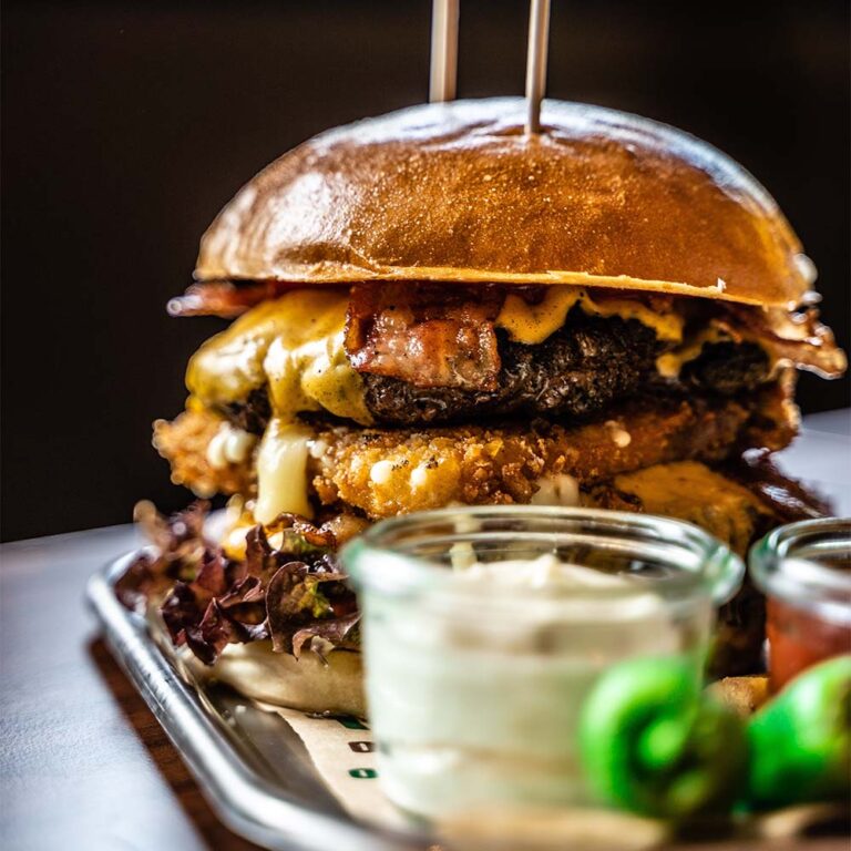 A metal tray with glass sauce jars and a large burger filled with a beef patty, melted cheese, bacon, lettuce, all held together with two skewers piercing the glazed bun.