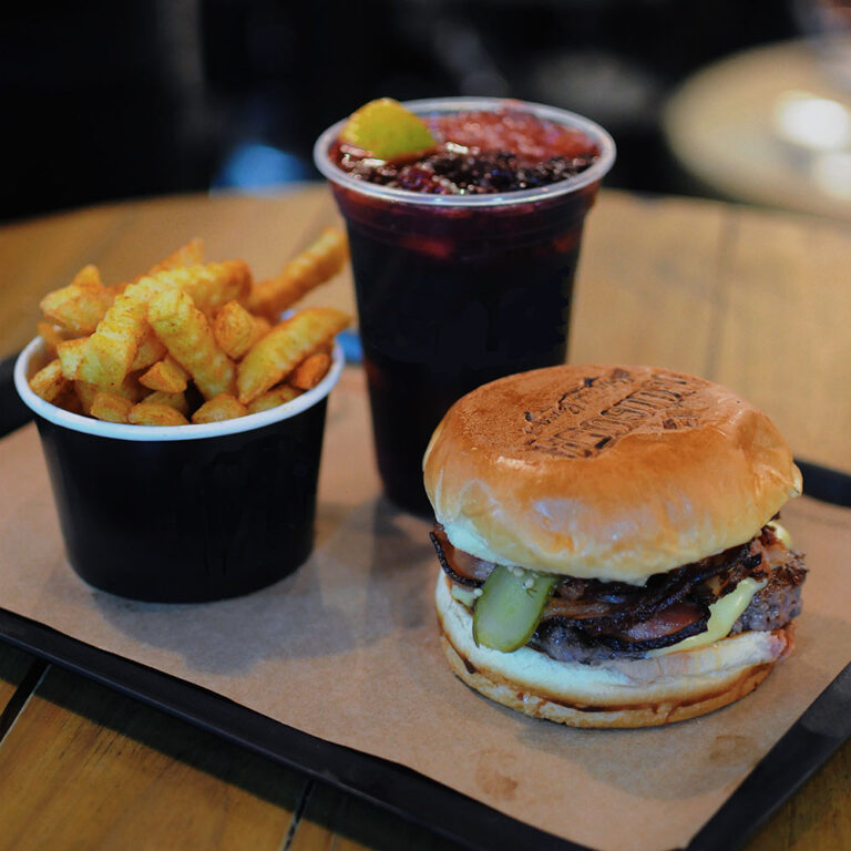 A round wooden table with a black tray with brown paper liner holding a burger, black paper cup of hot chips, and a plastic cup of soft drink.