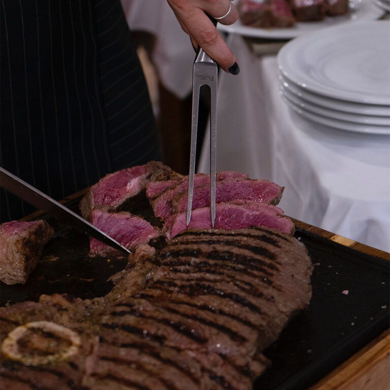 A very large, rare steak on a cutting board being sliced with a carving fork and knife by a white woman wearing silver rings and black nail polish, with a stack of white plates in the background.