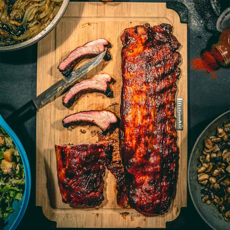 A wooden cutting board with two slabs of glistening barbeque ribs on it, one of the ribs has been cut into pieces with a knife, surrounded by bowls of salad.